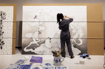 An artist is working on a large mural featuring female figures.
